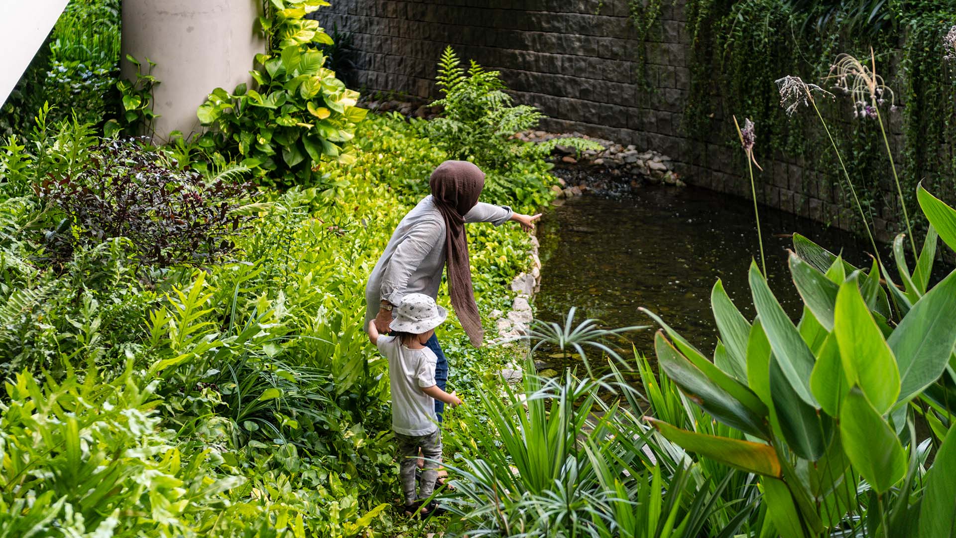 Scorcio della vegetazione rigogliosa ai margini del biotopo artificiale che attraversa l’edificio. Una donna con un bambino osserva la fauna acquatica, sottolineando come gli spazi verdi favoriscano l’interazione tra persone e ambiente | ©Lim Wei Xiang, Henning Larsen Architects